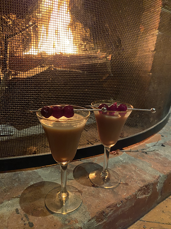 Two sherry glasses garnished with cherries sit on a brick hearth in front of a fire.