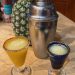 Two shotglasses of The Smoky Pineapple sit in front of a pineapple and cocktail shaker.